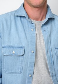 Camisa de mezclilla azul clara con cuello, dos bolsillos en el pecho y botones blancos, superpuesta sobre una camiseta gris de cuello redondo. Textura del tejido de algodón visible.