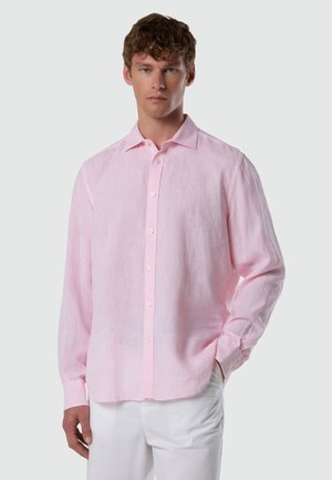 Junger Mann mit lockigem Haar, der ein hellrosa langärmliges Hemd mit Knöpfen und eine weiße Hose vor einem schlichten Hintergrund trägt.