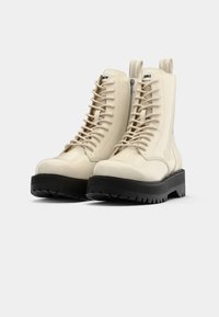 Botines de charol beige con cordones, cierre de cremallera y suela de goma negra texturizada. Presentan lengüetas y acabados contrastantes.