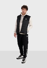 Chaleco de plumas negro con parte delantera acolchada, llevado sobre una sudadera crema. Combinado con pantalones cargo negros y zapatillas con detalles en blanco. Logos visibles.