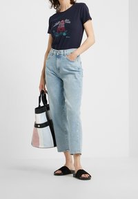 Kobieta w granatowej koszulce z wzorem latarni morskiej, jasnoniebieskich dżinsach, czarnych klapkach, trzymająca dużą torbę w paski.