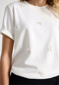 Hvid kortærmet t-shirt lavet af blødt stof, med flere teksturerede, cremefarvede sløjfeapplikationer over forsiden.