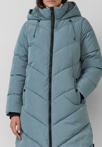 Frau, die einen hellblauen, gesteppten Wintermantel mit Kapuze, schwarzem Reißverschluss und Taschen hat.