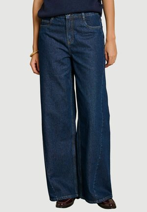 Person, die dunkelblaue Jeans mit weitem Bein trägt, kombiniert mit dunkelbraunen Schuhen und einem dunkelblauen Oberteil vor einfachem Hintergrund.