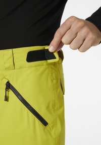 Kirkad kollased püksid tekstuurse viimistlusega, millel on lukuga tasku ja must Velcro rihm vöökohtade reguleerimiseks.