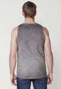 Sivina brezrokavna majica z teksturiranim, degradiranim vzorcem. Ima okrogel izrez in ohlapen kroj, v kombinaciji z modrimi jeans hlačami.