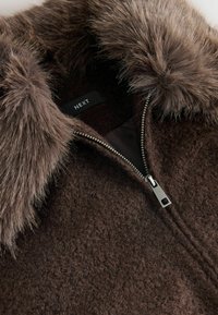 Chaqueta de forro polar marrón con cuello de piel sintética, cierre de cremallera con herrajes plateados y tejido texturizado. La etiqueta dice "NEXT."