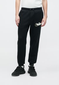 Melnas sporta bikses no mīksta auduma ar ribotu jostu, elastīgām aprocēm un baltu Nike logotipu izšūtu uz kreisās augšstilba daļas.