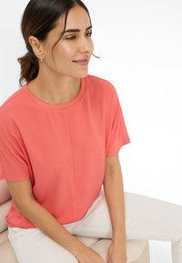 Coral short-sleeve top made of smooth fabric with a round neckline and subtle design details, paired with white trousers.