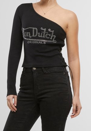 Femme portant un top noir à une manche avec le texte "Von Dutch Originals" et un jean noir taille haute, montrée des épaules aux hanches.
