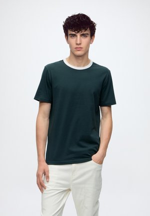 Ung mand med mørkt krøllet hår iført en mørkegrøn t-shirt med hvid krave og lyse bukser mod en ensfarvet baggrund.