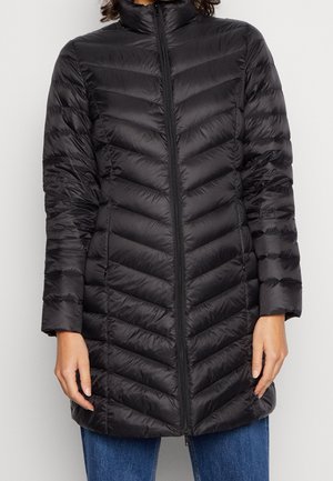 Kvinde iført en sort, quiltet, lang dunjakke med høj krave og lynlås foran, kombineret med blå jeans.