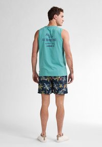 Světle tyrkysové bezrukávové tričko s grafikou "Key West" na zádech, spárované s námořnickými šortkami se barevným vzorem palmových stromů. Pohodlné boty.