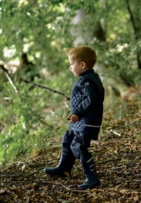 Niño con chaqueta azul marino acolchada y pantalones a juego, de pie sobre suelo cubierto de hojas, sosteniendo un palo, con botas de goma azules.