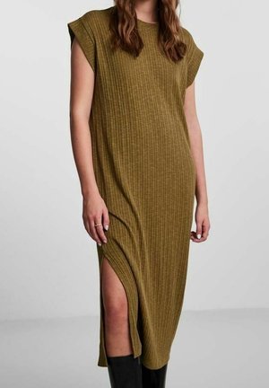 Mujer vistiendo un vestido midi de punto verde oliva sin mangas con abertura lateral y botas negras hasta la rodilla, de pie frente a un fondo gris.