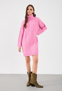 Svijetlo ružičasta pletena haljina s turtleneck izrezom, s cable uzorcima, dugim rukavima i rebrastim rubom. Uparena s maslinasto zelenim kaubojskim čizmama.