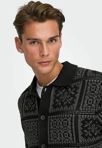Svart och grå mönstrad stickad cardigan med en texturerad yta, knäppning fram och krage. Har geometriska och blommiga mönster.