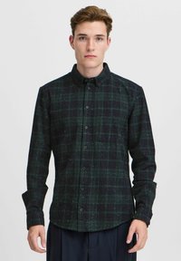 Cămașă cu carouri verzi și negre, cu butoni, un buzunar pe piept, mâneci lungi și textură presărată. Material din bumbac, design casual.