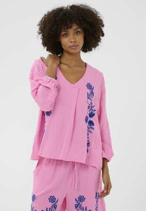 Vrouw met krullend haar die een loszittende roze outfit met blauwe bloemenborduursels draagt, met één hand op haar schouder staand tegen een effen achtergrond.