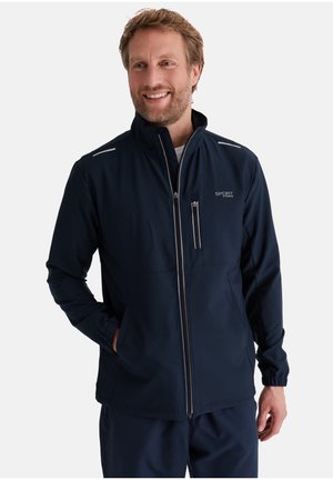 Lächelnder Mann, der eine marineblaue Sportjacke mit Reißverschluss und dem "SPORT"-Logo trägt, steht mit einer Hand in der Tasche vor einem einfarbigen Hintergrund.