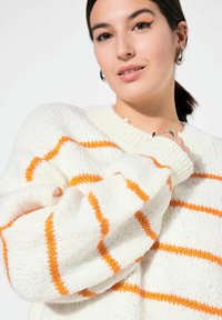 Weißer gestrickter Pullover mit orangefarbenen Streifen, im lässigen Schnitt und mit gerippten Bündchen. Weiche Textur, sichtbare Nähte im gesamten Pullover.