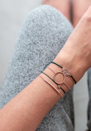 Dünne, gestapelte Armbänder aus Roségold und grünem Schnur mit einem Blätteranhänger, einem Herz und einem gedrehten Ring-Design, vor einem strukturierten grauen Hintergrund.
