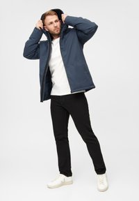 En mand i en marineblå hættetrøje justerer sin hætte, iført en hvid skjorte og sorte jeans, stående mod en ensfarvet lys baggrund.