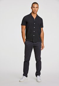 Μαύρο κοντομάνικο πουκάμισο με γιακά, άσπρα κουμπιά και μαλακή υφή, συνδυασμένο με σκούρο παντελόνι και άσπρα αθλητικά παπούτσια. Απλή, καθημερινή σχεδίαση.