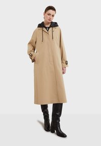 Beige trenchcoat med svart huva, justerbara dragsnören, sidofickor och spännband på ärmarna. Slät tygfinish.