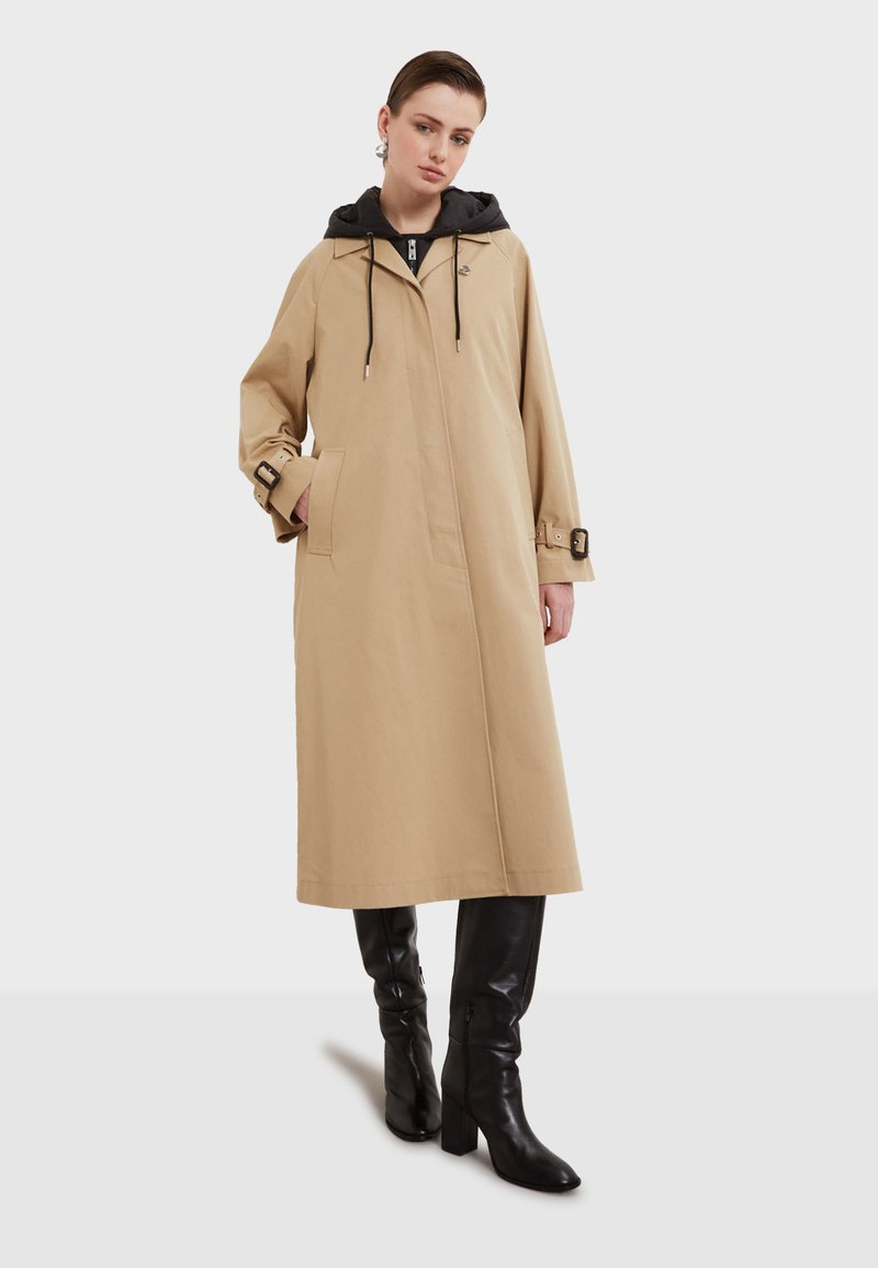 Beige trenchcoat med svart huva, justerbara dragsnören, sidofickor och spännband på ärmarna. Slät tygfinish.