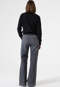 Un pull noir texturé associé à un jean large taille haute gris foncé avec des poches arrière et des coutures discrètes, sur un fond blanc uni.