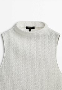 Haut blanc sans manches texturé avec un col rond haut et une étiquette noire à l’intérieur du col.