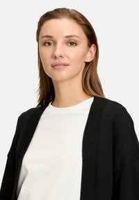 Svart kofta med ribbade detaljer över en vit skjorta. Slät textur, öppen framsidan design och avslappnad passform. Enkelt, mångsidigt stil.