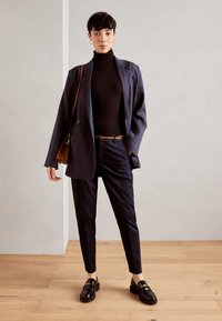 Marineblazer über einem schwarzen Rollkragenpullover, kombiniert mit maßgeschneiderten marineblauen Hosen und schwarzen Mokassins. Inklusive braunem Gürtel und Schultertasche.