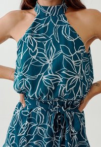 Teal halterneck-blus med vitt blommönster, lös passform, rynkad midja och knytdetalj. Mjuk tygtextur med en slät finish.