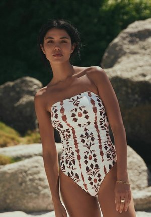 Femeie stând în aer liber, purtând un costum de baie alb fără bretele, cu modele abstracte maro și negre, cu stânci și verdeață în fundal.