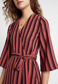 Robe portefeuille rayée rouge et noire avec ceinture à nouer, dotée d'un col en V et de manches évasées, réalisée en tissu doux et léger.