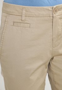 Beige chinos med bälteshällor, en liten framficka, knappstängning, bar tillsammans med en ljusblå skjorta instoppad.