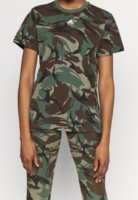 Tričko s maskáčovým vzorem v zelené a hnědé barvě, vyrobené z bavlny; má kulatý výstřih a malé bílé logo Adidas na hrudi.