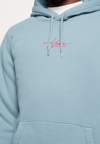 Ljusblå hoodie i mjukt material med en rosa broderad logotyp på bröstet, justerbara dragkedjor och en framficka.