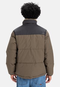 Pufferjack in bruin en zwart, met een hoge kraag, horizontaal gestikt ontwerp en een zachte, gestructureerde afwerking. Verstelbare zoom.