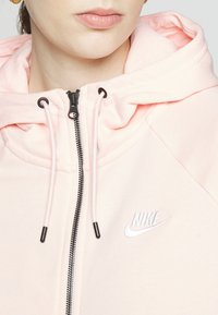Ljusrosa zip-up hoodie med svart dragkedja, justerbara snoddar och broderad vit Nike-logga. Mjuk tyg och avslappnad design.