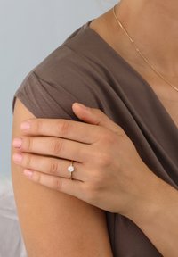 Gold ring with a round, clear gemstone, set on a slim band. A hand rests on a shoulder, featuring a smooth, muted fabric.