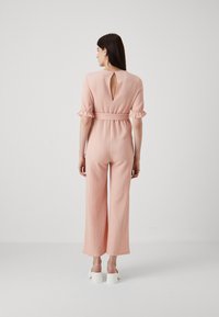 Rosa teksturert jumpsuit med korte volangermer, nøkkelhulutskjæring bak, belted midje og vide bukser. Brukt med hvite blokkhelsesko.