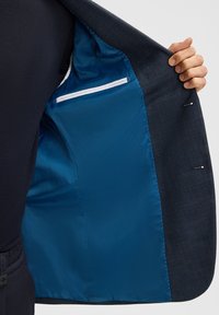 Hand die een donkerblauwe blazer vasthoudt en de gladde blauwe voering en binnenzak met witte rand laat zien.