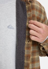 Braun-grün kariertes Flanellhemd mit einem grauen, mit Fleece gefütterten Kragen, getragen über einem hellgrauen Shirt mit kleinem Logo.