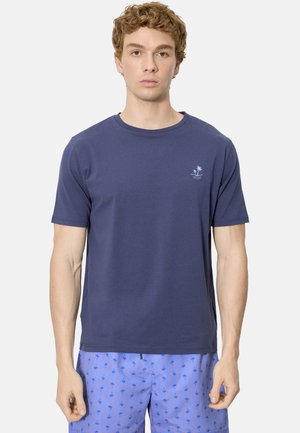 Joven con camiseta azul marino con un pequeño logo de palmera y pantalones cortos azules con estampado, posando frente a un fondo blanco liso.