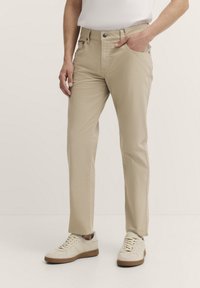 Beige Baumwollhose mit geradem Schnitt, Fronttaschen und Gürtelschlaufen, getragen mit hellen Sneakern vor einem neutralen Hintergrund.