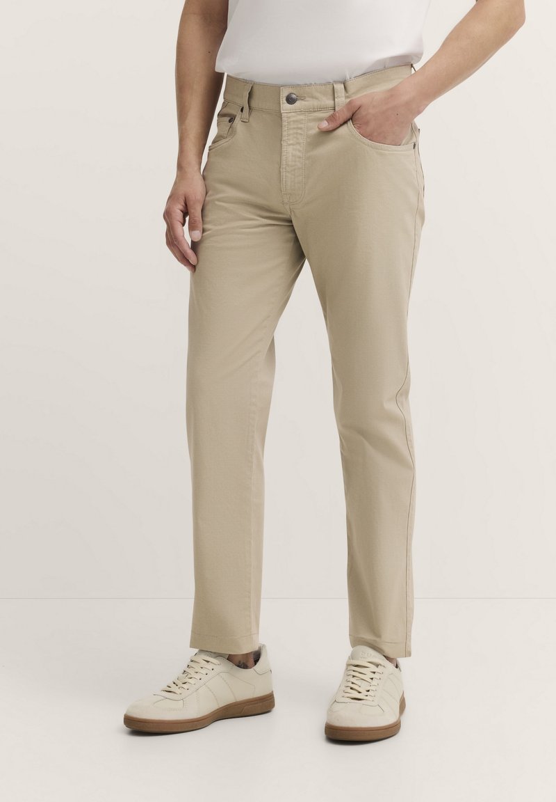 Beige Baumwollhose mit geradem Schnitt, Fronttaschen und Gürtelschlaufen, getragen mit hellen Sneakern vor einem neutralen Hintergrund.
