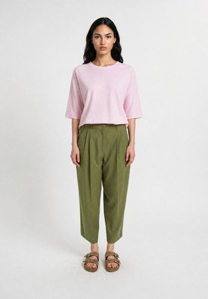 Frau mit langen dunklen Haaren, die ein lockeres rosa Hemd, olivgrüne, gekürzte Hosen und braune Sandalen trägt, steht vor einem einfachen hellen Hintergrund.